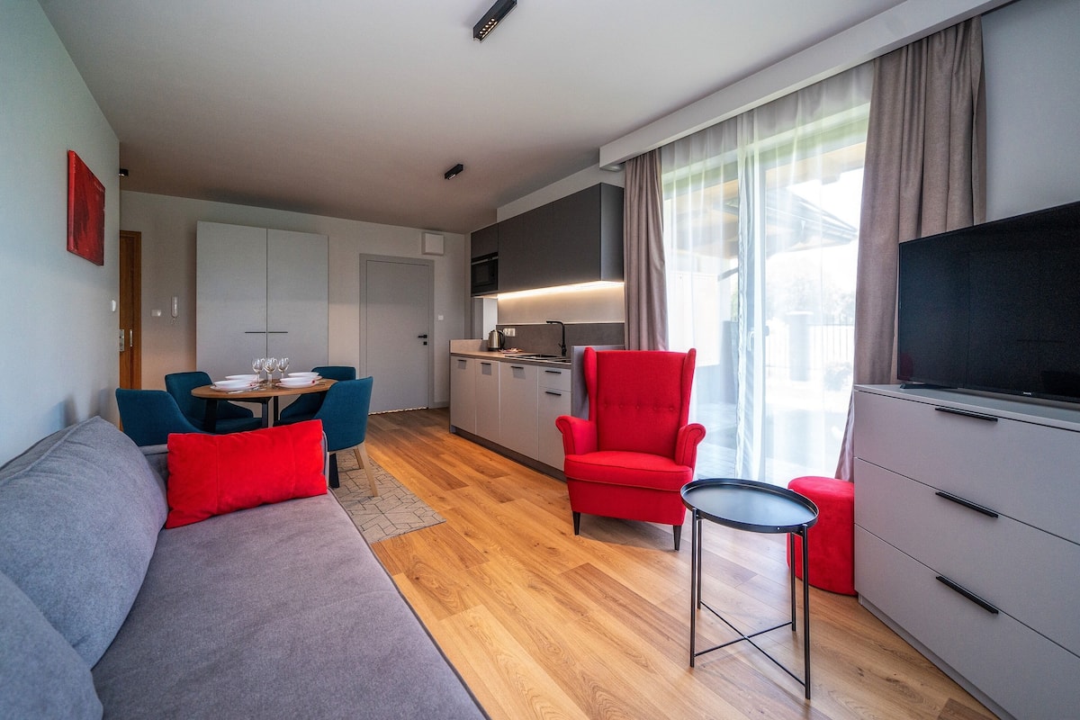 Uma sala de estar moderna com um sofá cinzento, uma poltrona vermelha e uma mesa pequena. Há uma área de cozinha com uma mesa de jantar e cadeiras, e uma TV sobre um armário.