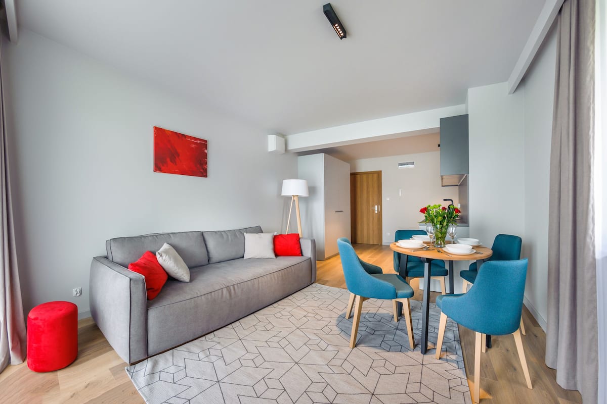 Uma sala de estar moderna com um sofá cinza, almofadas vermelhas e brancas, um banquinho vermelho e um tapete com padrão geométrico. Há uma área de jantar com uma mesa redonda, cadeiras azuis e um vaso de flores.