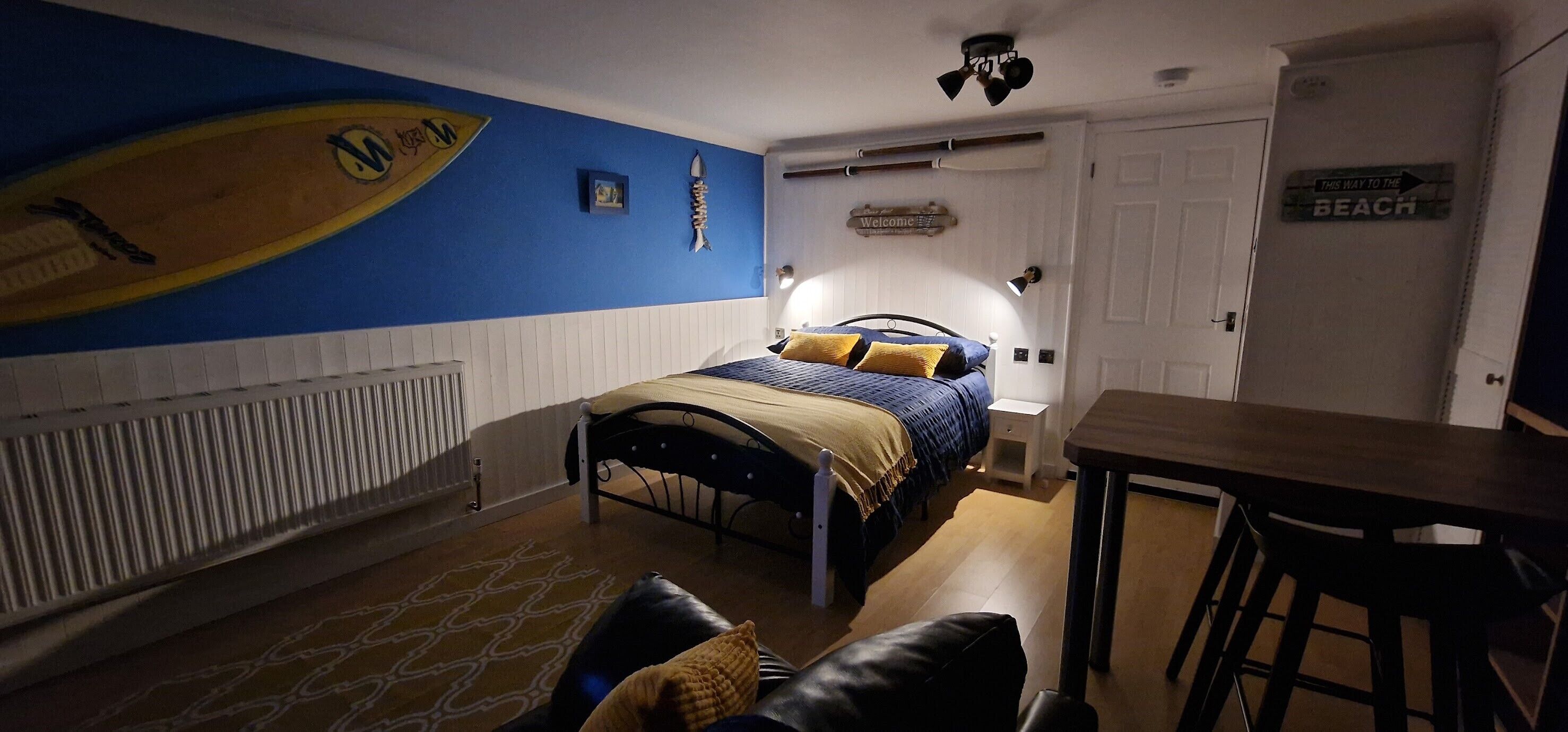 A bedroom with a bed, a desk, a chair, a radiator, and a wall with a surfboard and a 'Welcome' sign.