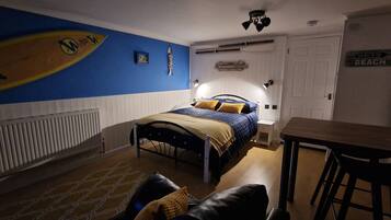 A bedroom with a bed, a desk, a chair, a radiator, and a wall with a surfboard and a 'Welcome' sign.