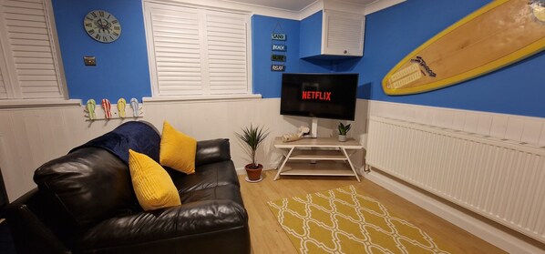 A living room with a black leather sofa, yellow cushions, a TV on a stand, a surfboard, and a clock on the wall.