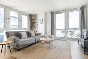 A modern living room with a grey sofa, a round coffee table, and a large window with curtains. - The Bow Flats (London)