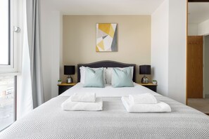 A hotel room with a bed, two bedside tables, a lamp, a wall art piece, and a view through a window. - The Bow Flats (London)