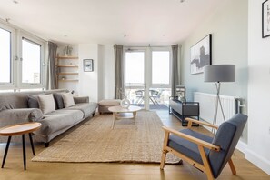 Un salon moderne avec un canapé gris, une table basse ronde en bois, un fauteuil bleu et une grande fenêtre avec des rideaux.