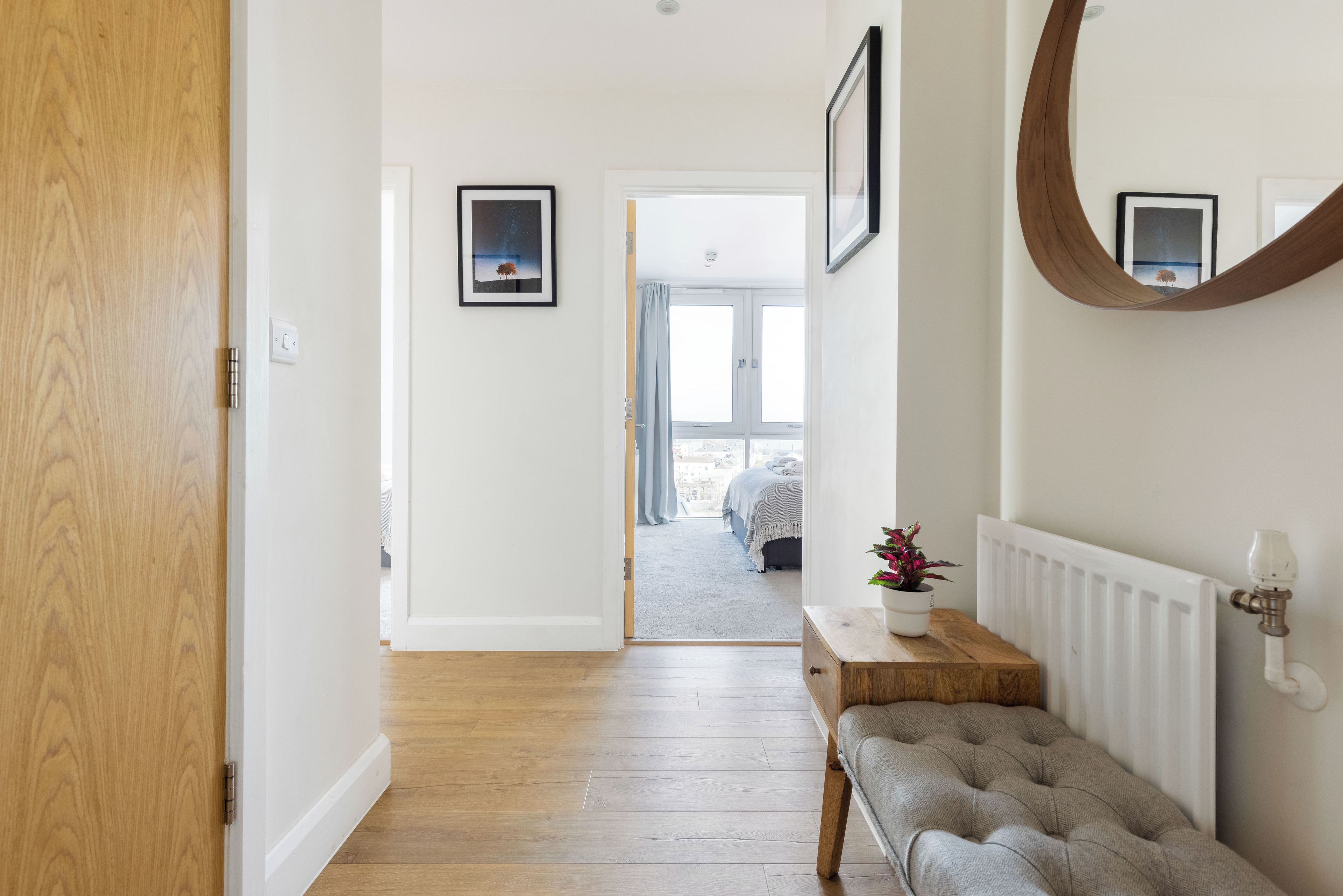 A hallway with a wooden floor, a bench, a radiator, and a door leading to a bedroom.