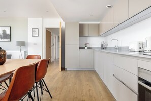 A modern kitchen with white cabinets, a wooden dining table with chairs, and a coffee maker. - The Bow Flats (London)