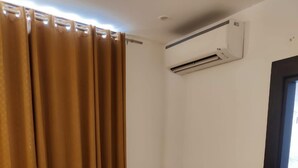 A room with a glass-top table, two wooden chairs with patterned cushions, a brown curtain, and an air conditioning unit on the wall. - Hotel Rivasa Regency (New Delhi)