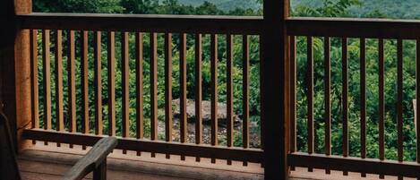 Une terrasse en bois, dotée d’une balustrade, offre une vue imprenable sur un paysage verdoyant et luxuriant, avec des montagnes à l’horizon.