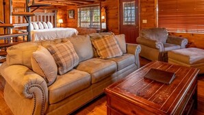A cozy log cabin interior with a sofa, armchair, and a wooden coffee table. A spiral staircase leads to a loft area with a bed. - Buffalo River Cabin w/ Hot Tub (Ponca)