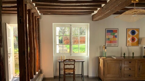 A room with a wooden ceiling, a window with a view of greenery, a table with a chair, and a cabinet with decorative items.