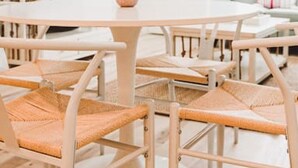 Ein modernes, helles Zimmer mit einem runden Tisch, umgeben von Korbstühlen, einer Topfpflanze und einem an der Wand montierten Fernseher.