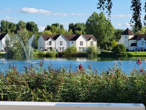 A lakeside view with houses, a fountain, and a person rowing a boat. - Luxurious South-Facing Lakeside Lodge (Cirencester)