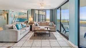 A spacious living room with a large window, a ceiling fan, and a view of the beach.