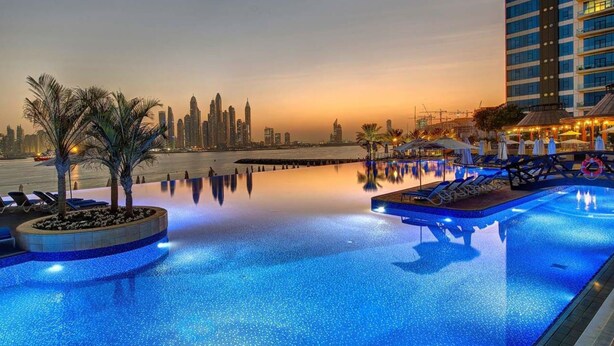 A poolside area with lounge chairs and palm trees, a city skyline in the background, and a high-rise building to the right. - Tiara - Luxury Palm Jumeirah (Dubai)