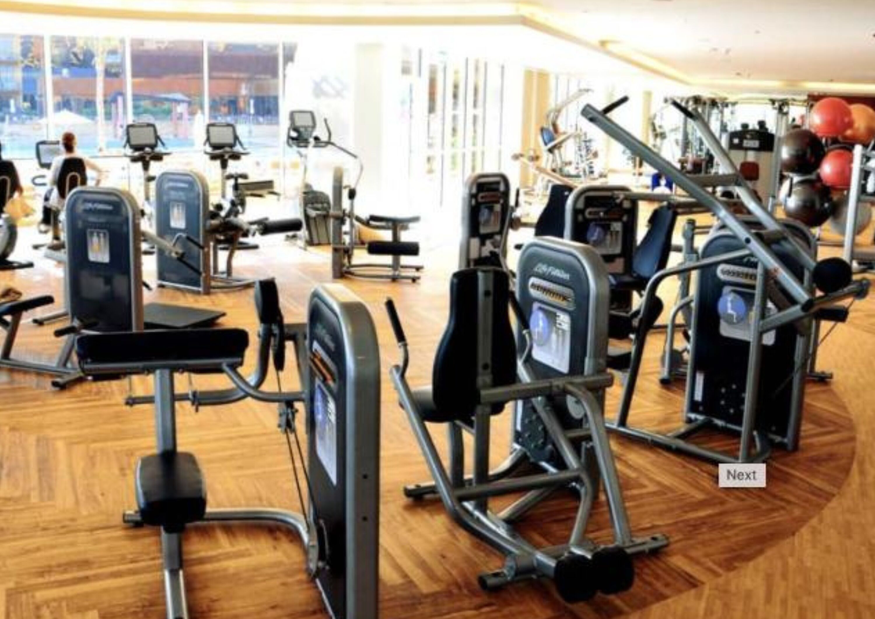 A gym with various exercise machines, including treadmills and weight machines, arranged on a wooden floor.