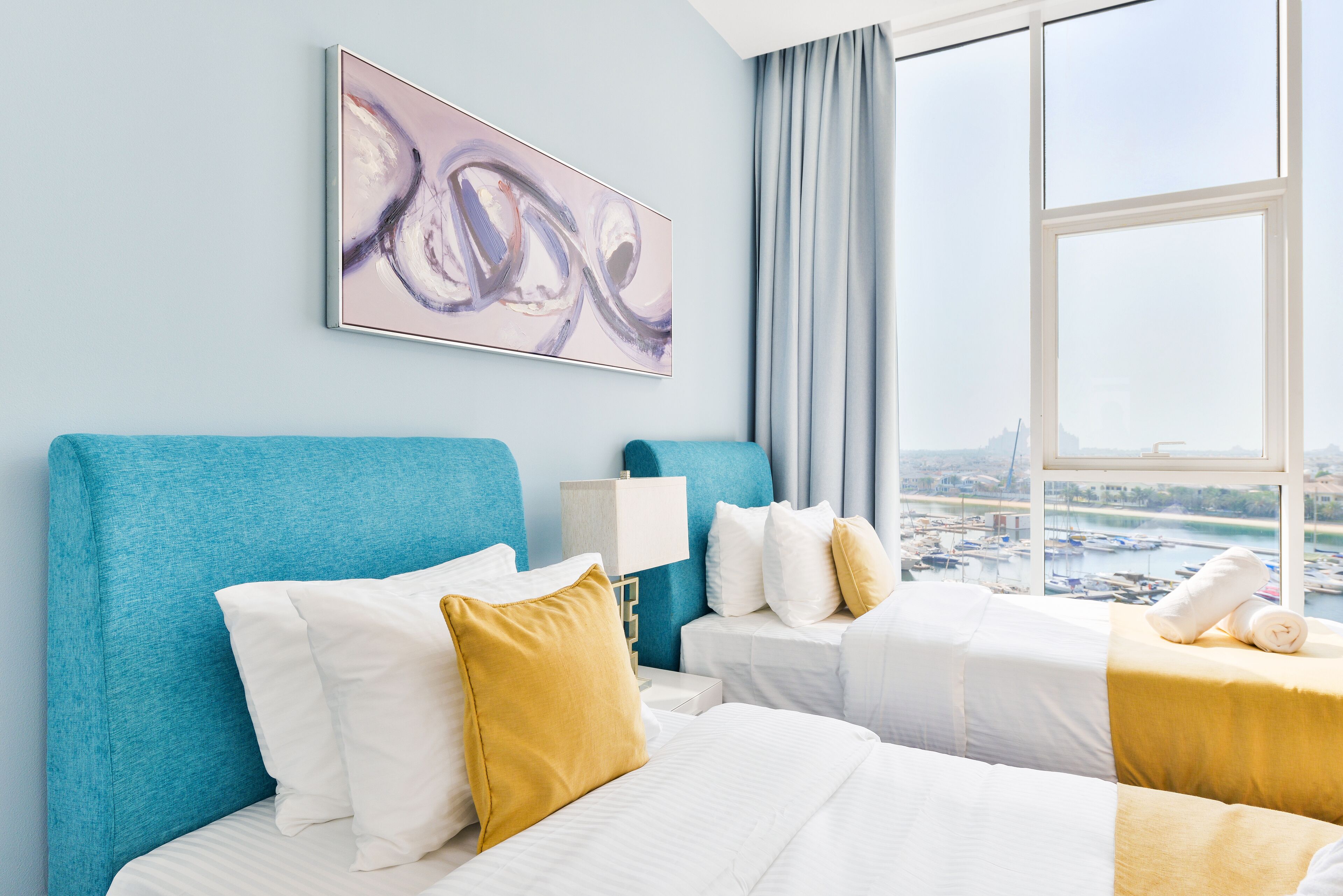 Une chambre d’hôtel avec une grande fenêtre, un lit avec une literie blanche et jaune, une tête de lit bleue et un tableau accroché au mur.