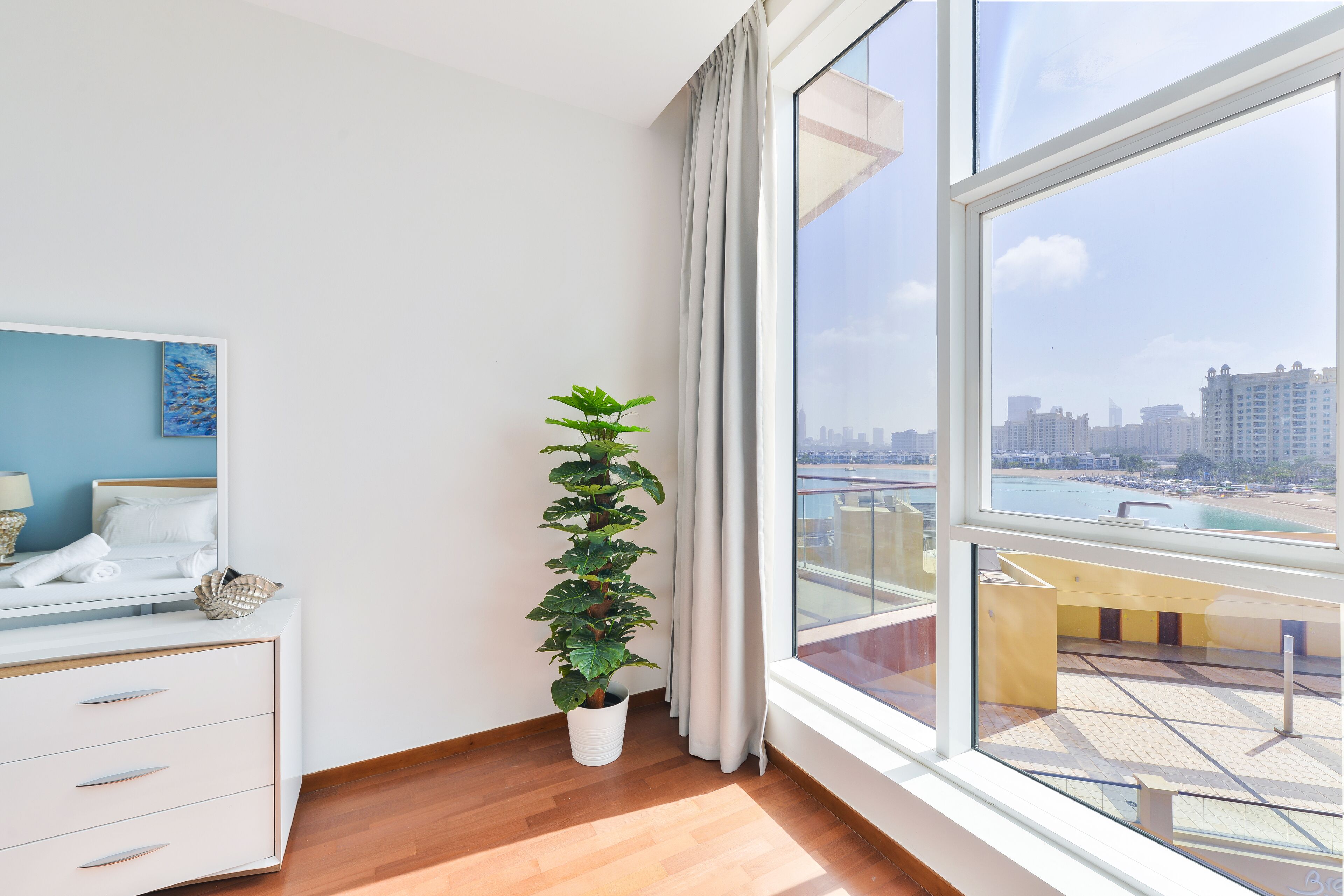 A modern hotel room with a large window, a potted plant, a bed with a blue headboard, and a white dresser.