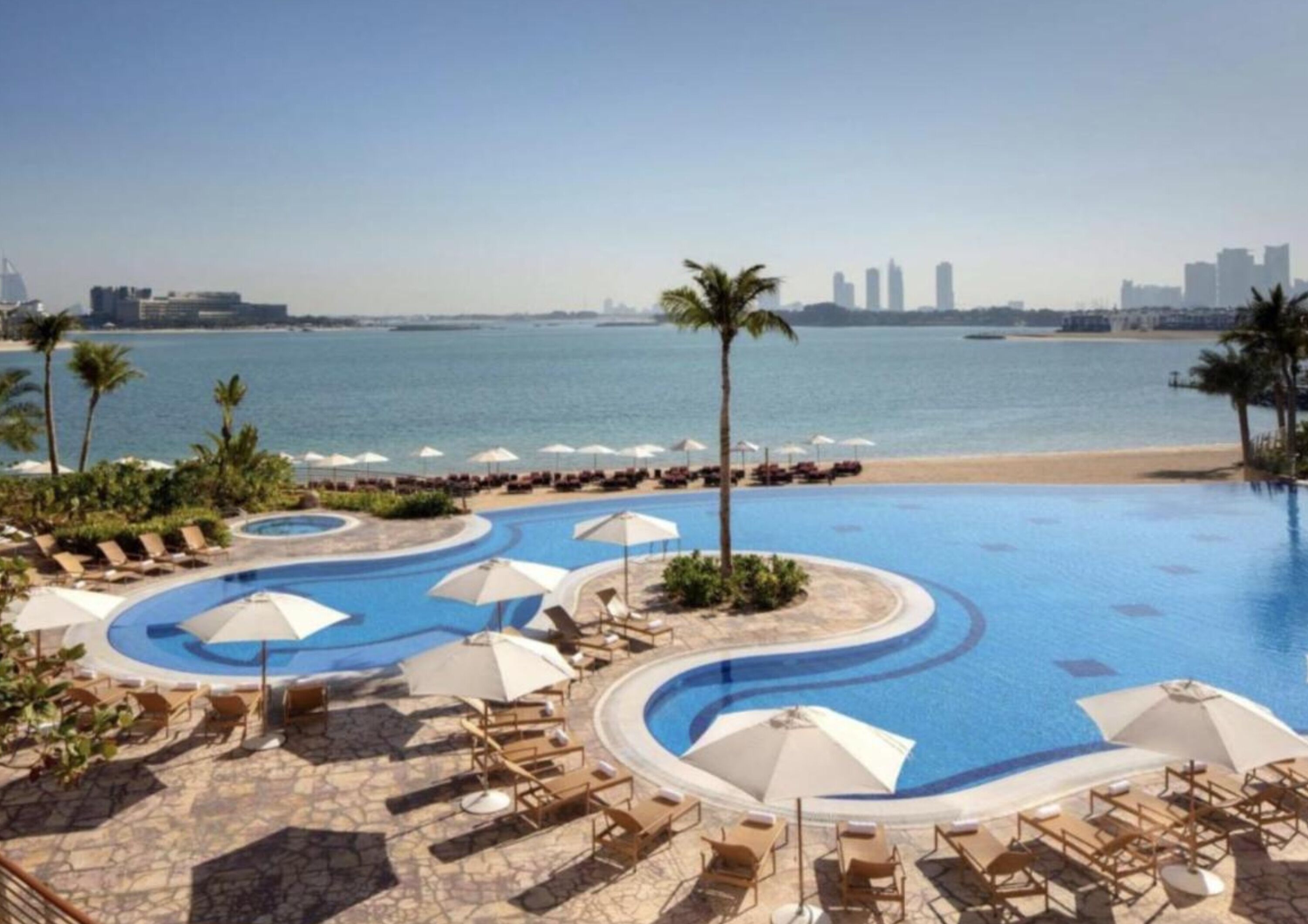 Een zwembad van een resort met een golvend ontwerp, omringd door ligbedden en parasols, met een strand en stadsachtergrond.