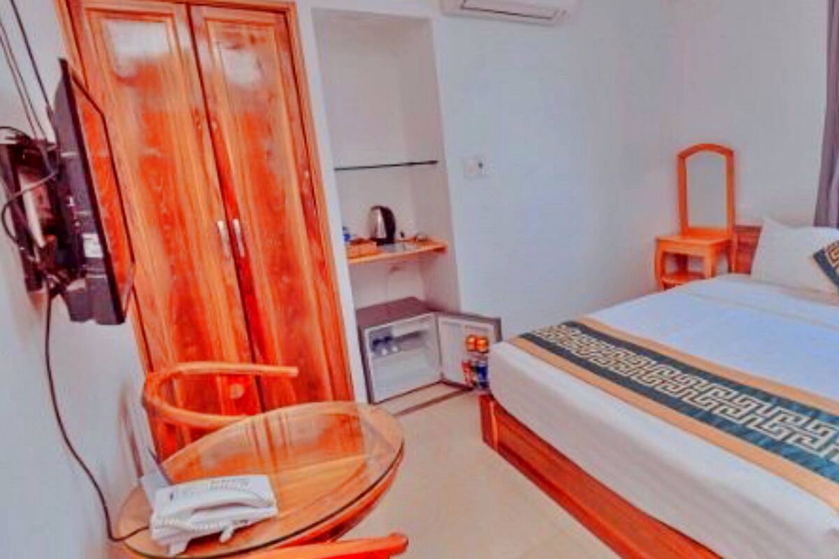 Une chambre d’hôtel avec un lit, une armoire en bois, une petite table avec une chaise et une fenêtre avec des rideaux.
