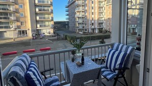 A balcony with two striped chairs, a small table with a vase and a bottle, and a view of a multi-story building with balconies.