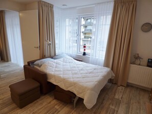 Ett hotellrum med en säng, ett nattduksbord, en lampa, ett fönster med gardiner och utsikt över byggnader utanför.