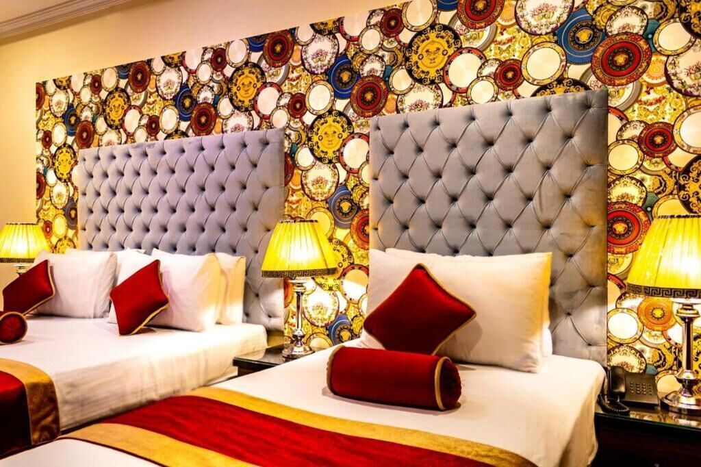 A hotel room with two beds, each with a tufted headboard, white bedding, red pillows, and a red throw blanket. There are two bedside lamps with yellow shades.