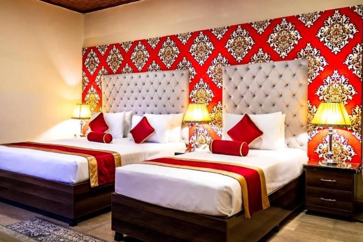 Habitación de hotel con dos camas, cada una con cabeceros capitonados, ropa de cama blanca y detalles en rojo. Hay lámparas de noche y un diseño decorativo en la pared.