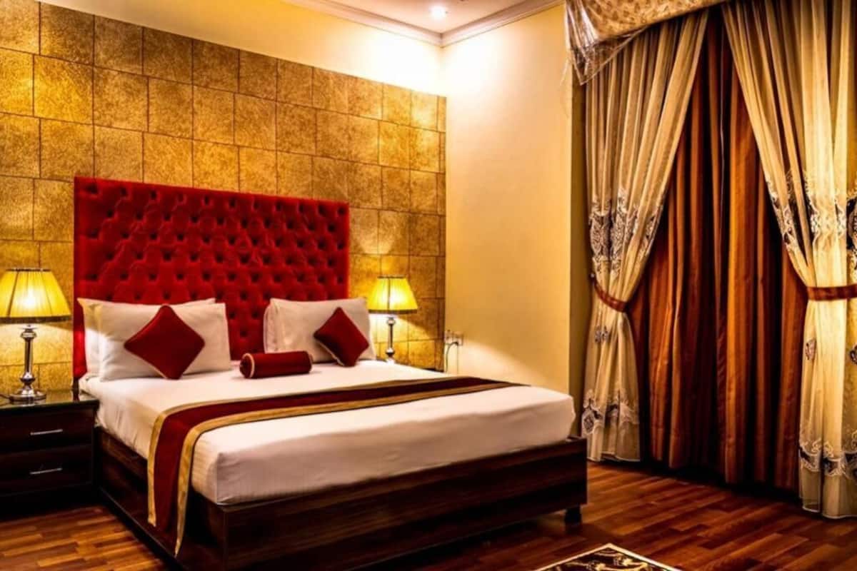 Habitación de hotel con un gran cabecero tapizado rojo, piso de madera y cama con sábanas blancas y almohadas rojas. Hay dos lámparas de noche y una ventana con cortinas.