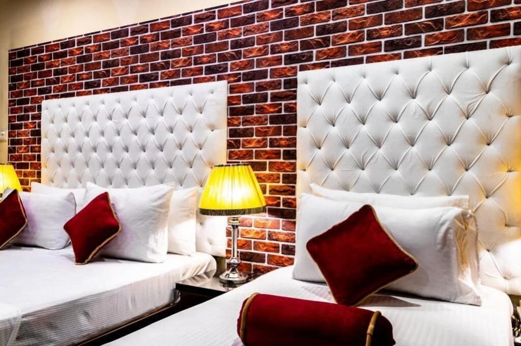 Una camera d'albergo con due letti, ognuno dotato di una testiera trapuntata, biancheria da letto bianca e cuscini rossi. Si nota una parete con decorazione a mattoni e una lampada gialla.
