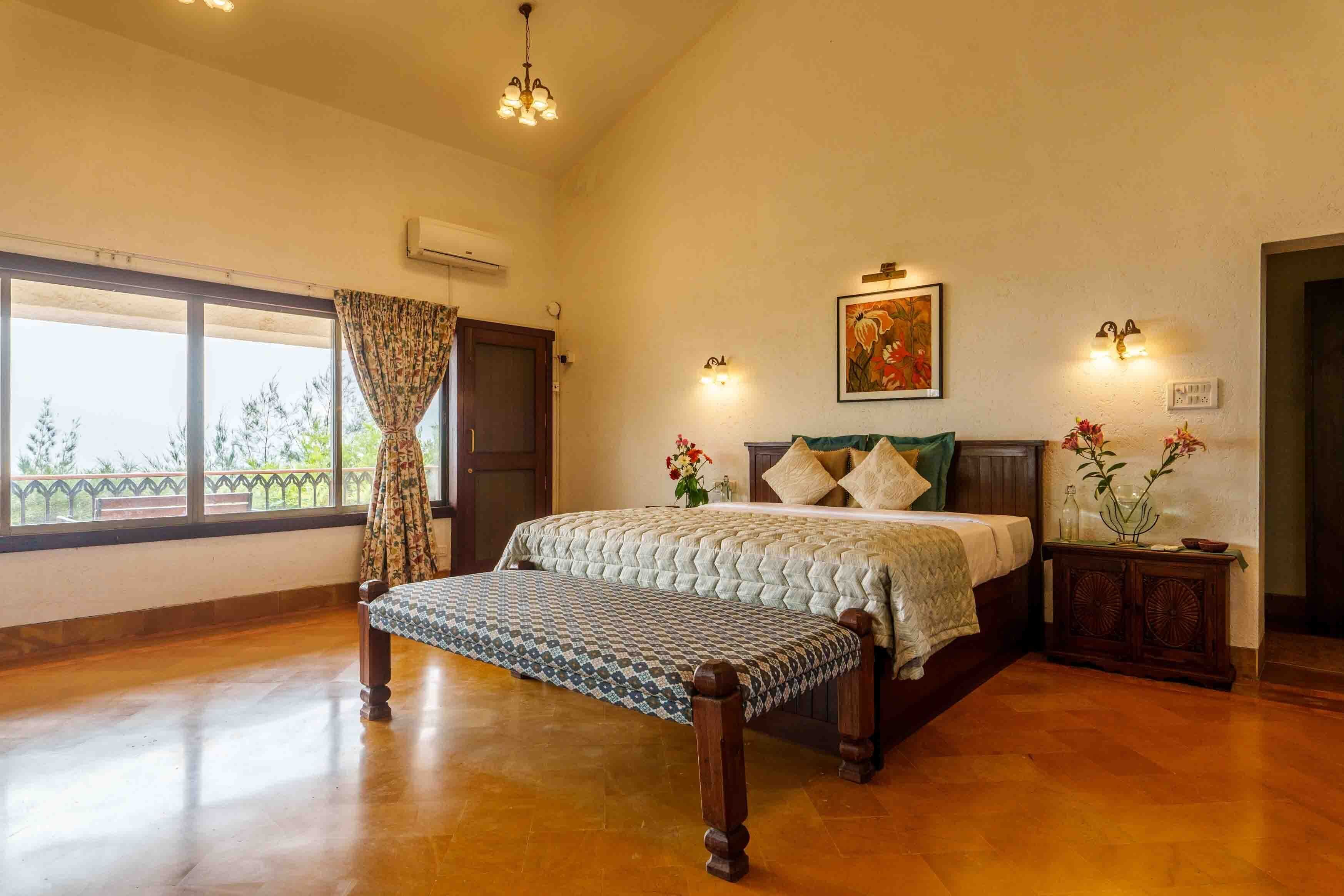 A bedroom with a bed, a bench, a nightstand, a window with curtains, and a painting on the wall.