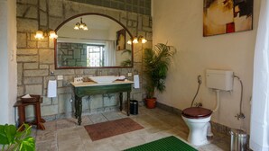 Een badkamer met een stenen muur, een houten wastafelmeubel, een wit toilet, een groene vloerkleed en een potplant.