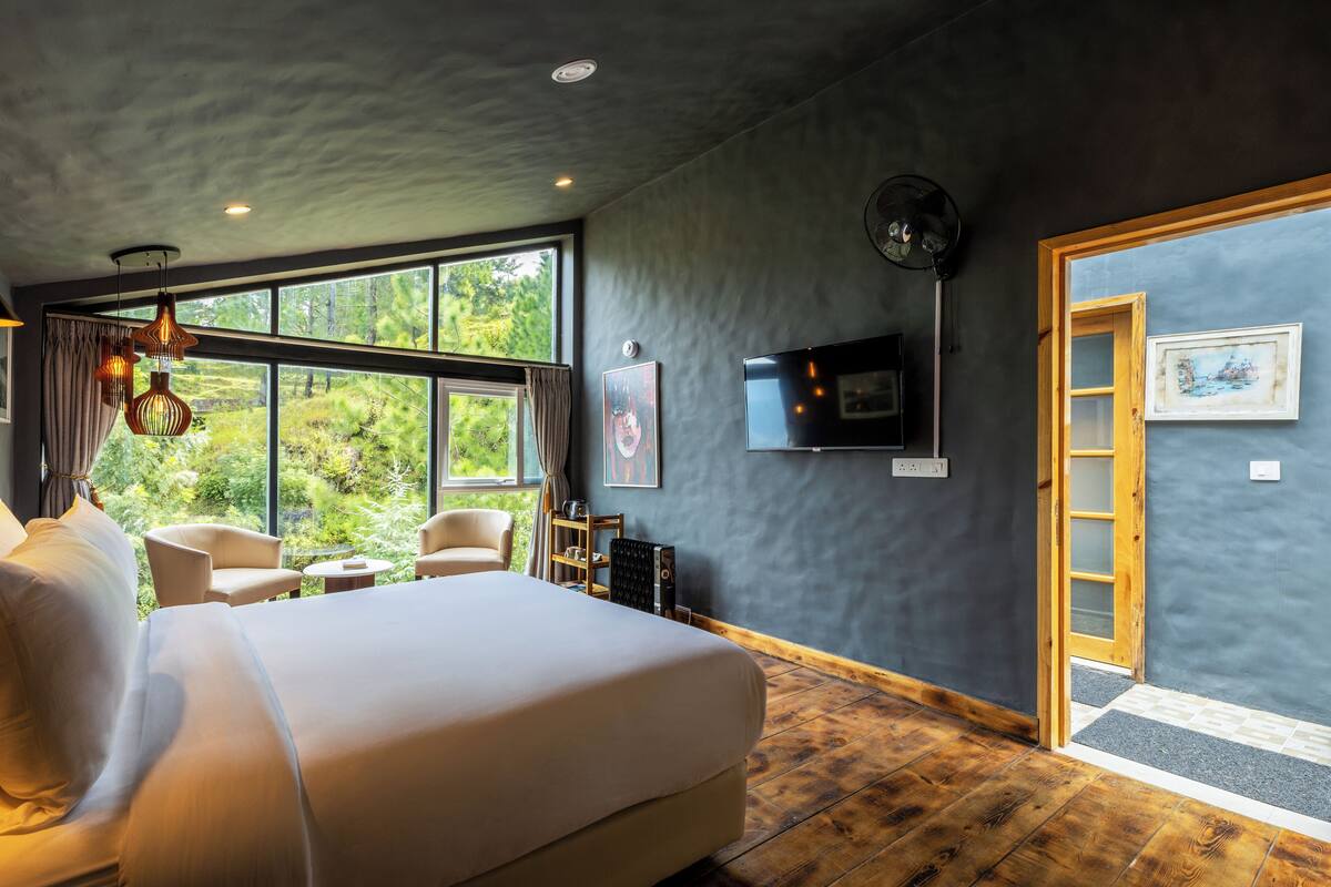 A bedroom with a large bed, two chairs, a small table, a TV, and a window with a view of greenery.