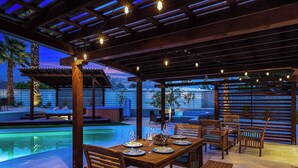 An outdoor dining area with a pool, wooden furniture, and string lights.