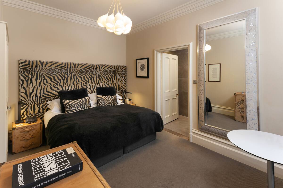 A bedroom with a zebra-patterned headboard, a bedside table with a book, a large mirror, and a small round table.