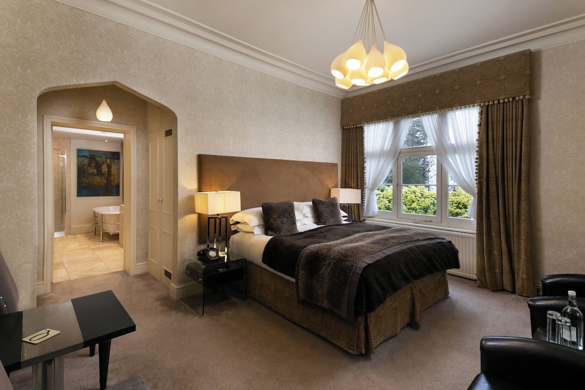A hotel room with a large bed, two bedside tables with lamps, a chair, and a view of greenery through the window.