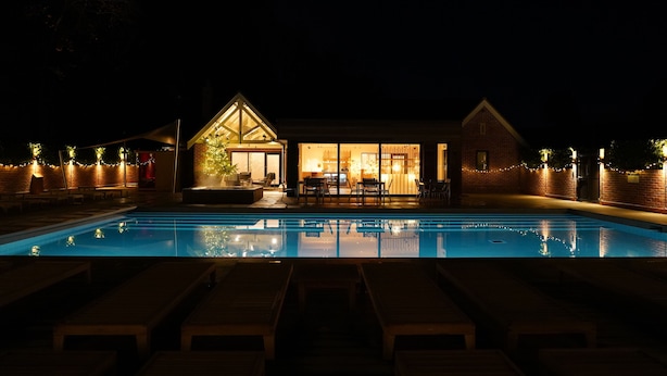 Vista nocturna de un área de piscina con sillones, una zona de bar y un edificio con amplios ventanales.
