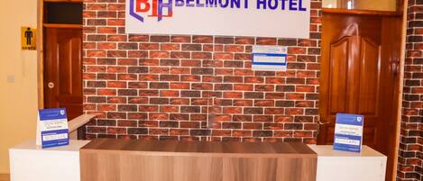 Un mostrador de recepción en el Hotel Belmont, con un cartel encima, una puerta de madera a la derecha y un letrero que indica un baño de hombres a la izquierda.