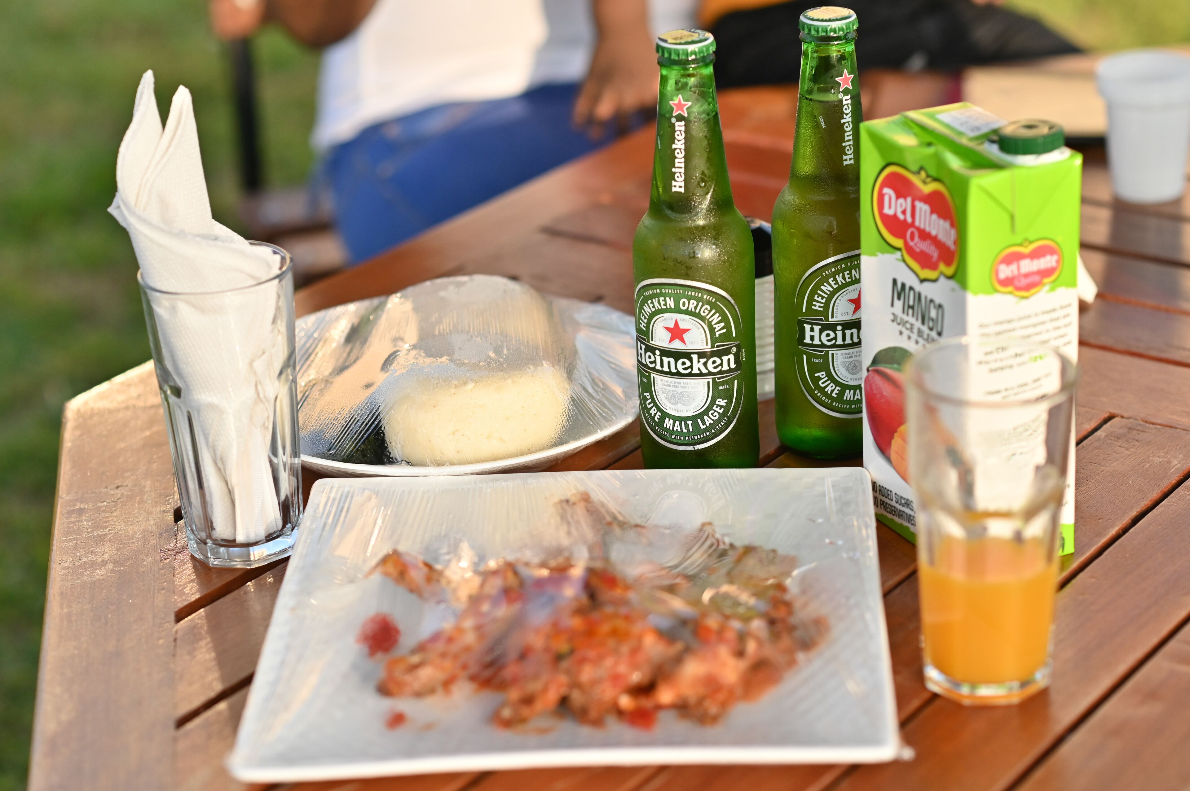 A picnic table with a plate of food, a glass of juice, and two bottles of Heineken beer.