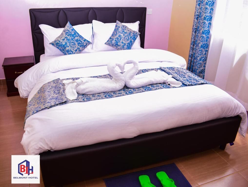 A hotel room with a black headboard, white bedding, and a blue floral bedspread. There are two pillows with a blue floral pattern, and a pair of green slippers on a blue mat.