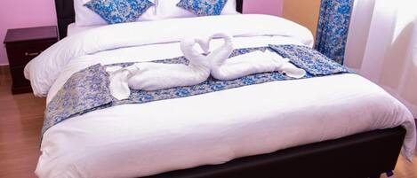 A hotel room with a black headboard, white bedding, and a blue floral bedspread. There are two pillows with a blue floral pattern, and a pair of green slippers on a blue mat.