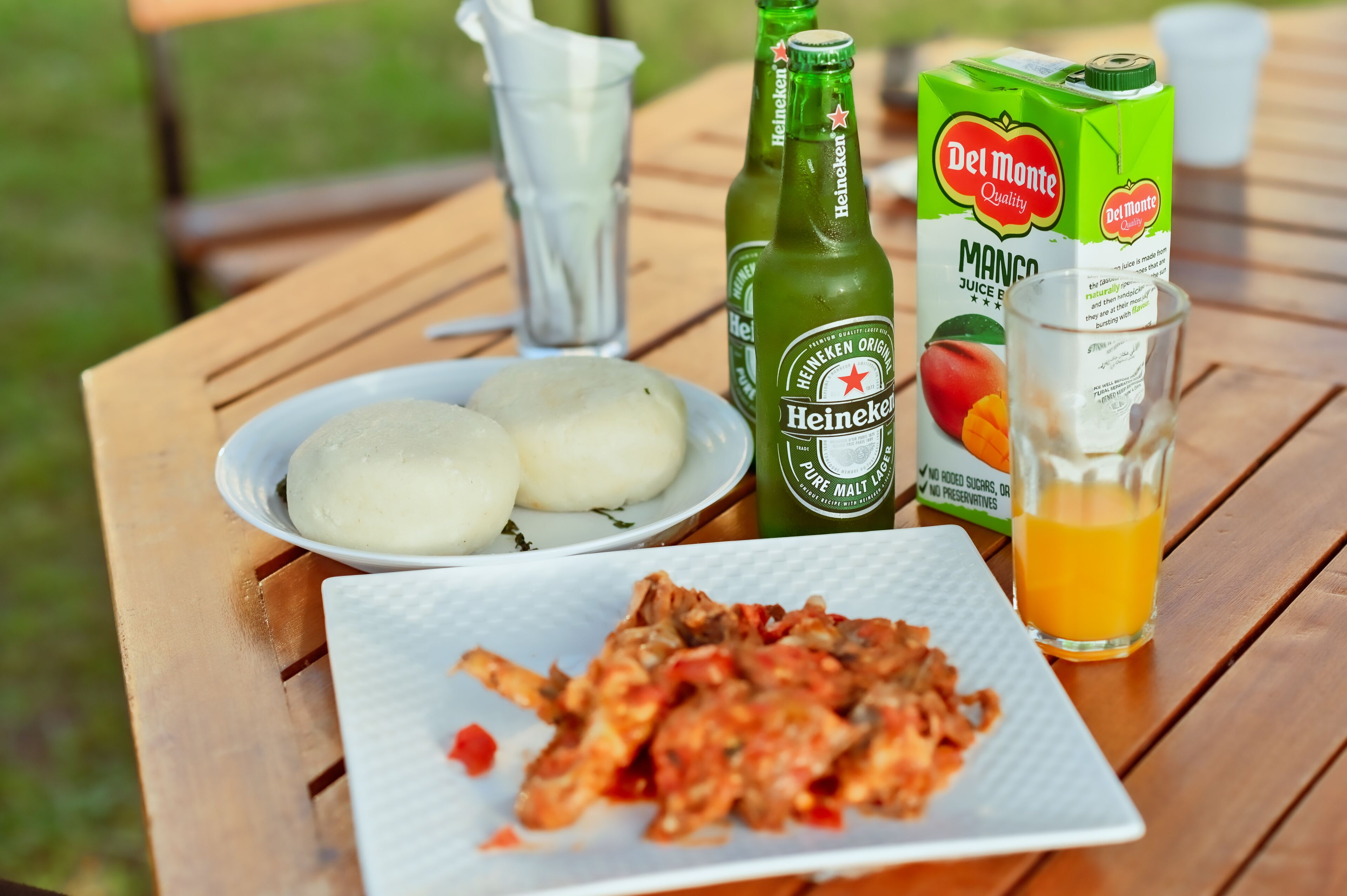 Una comida con albóndigas, un plato de comida frita, una botella de cerveza Heineken, un vaso de jugo y un cartón de jugo de mango sobre una mesa de madera.