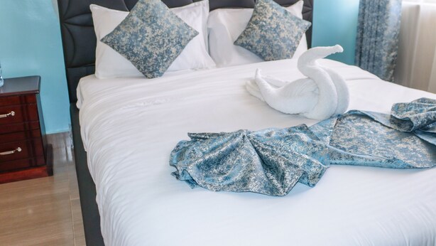A neatly made bed with a swan-shaped towel and a blue patterned blanket, a dark headboard, and a wooden nightstand.