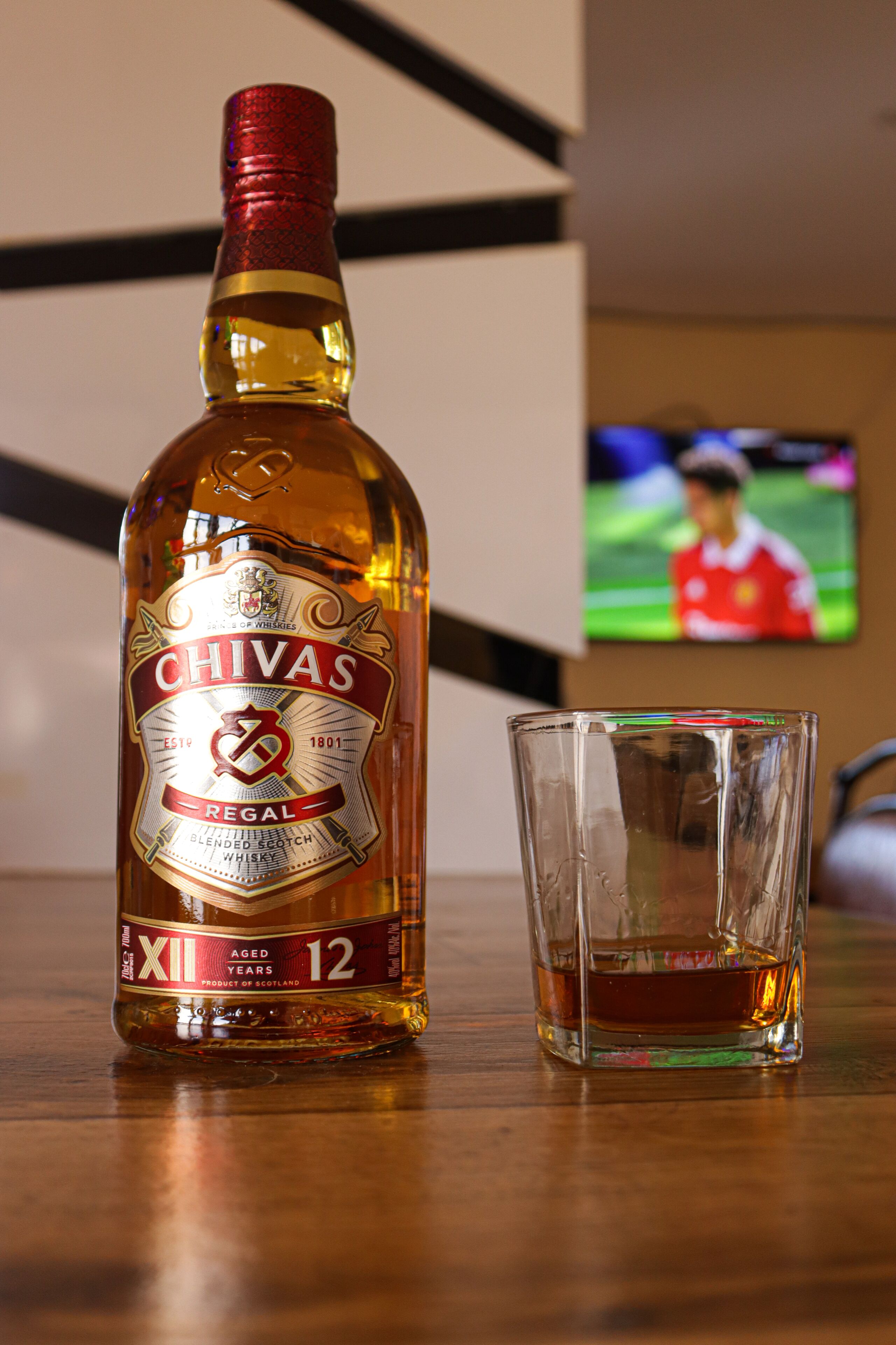 Una botella de whisky Chivas Regal y un vaso vacío sobre una mesa de madera.