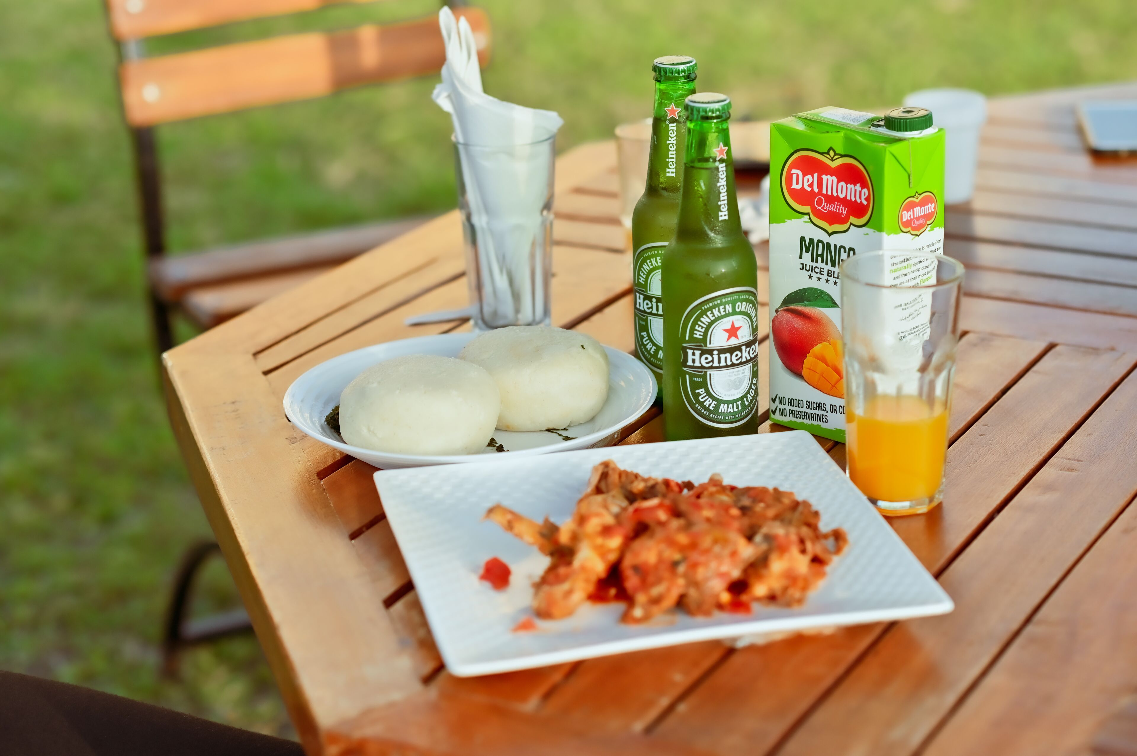 A wooden table with a plate of food, two bottles of beer, a glass of juice, and a carton of juice.