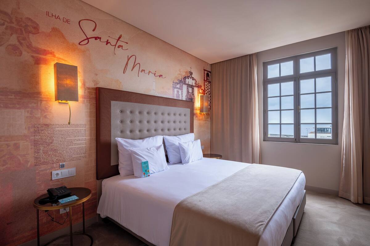 A hotel room with a large bed, a headboard, a nightstand, a wall-mounted lamp, and a window with curtains.