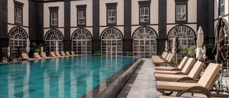 A pool area with lounge chairs and umbrellas, surrounded by a building with arched windows and a tiled floor.