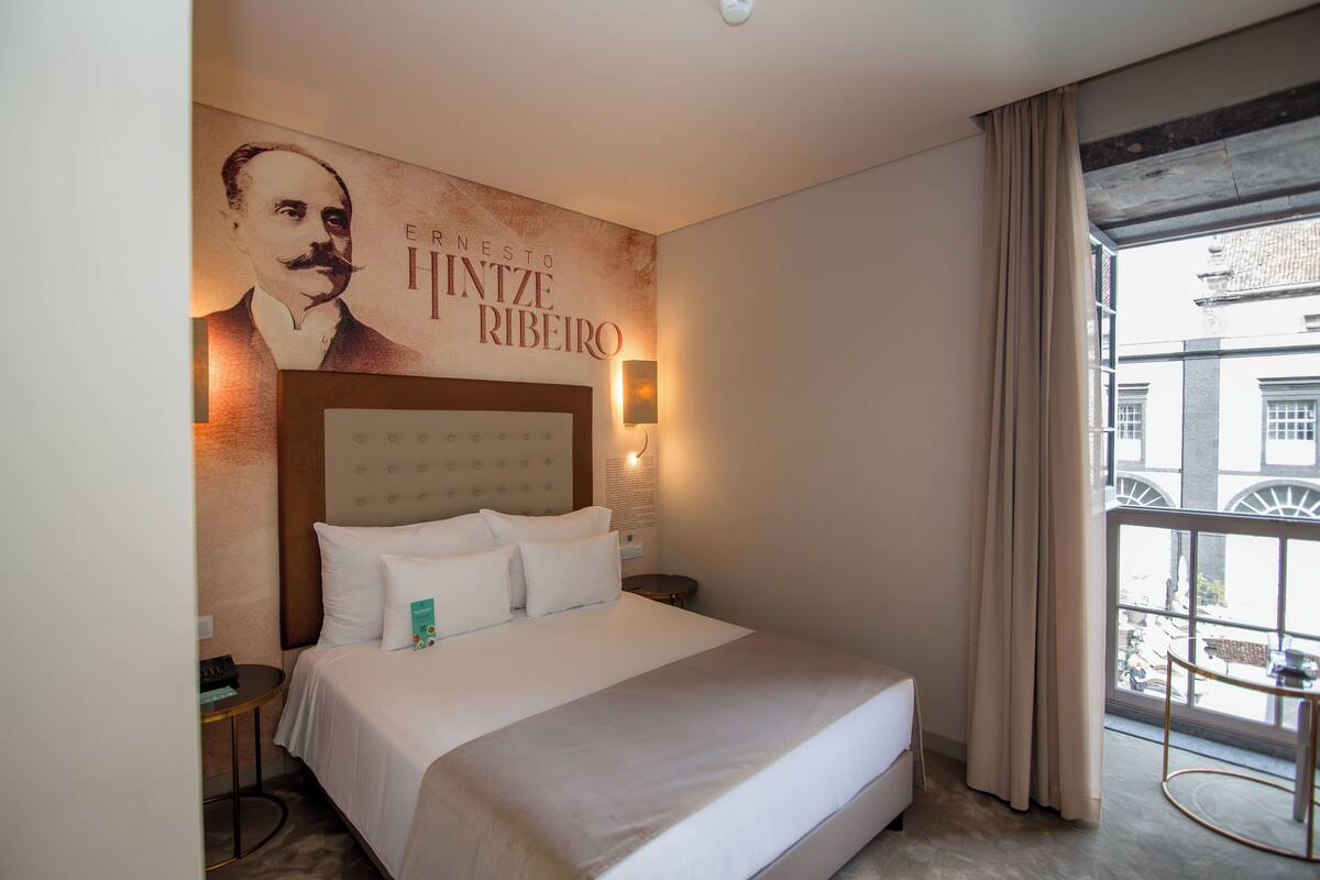 A hotel room with a large bed, a headboard, a nightstand, a wall-mounted lamp, and a window with curtains.