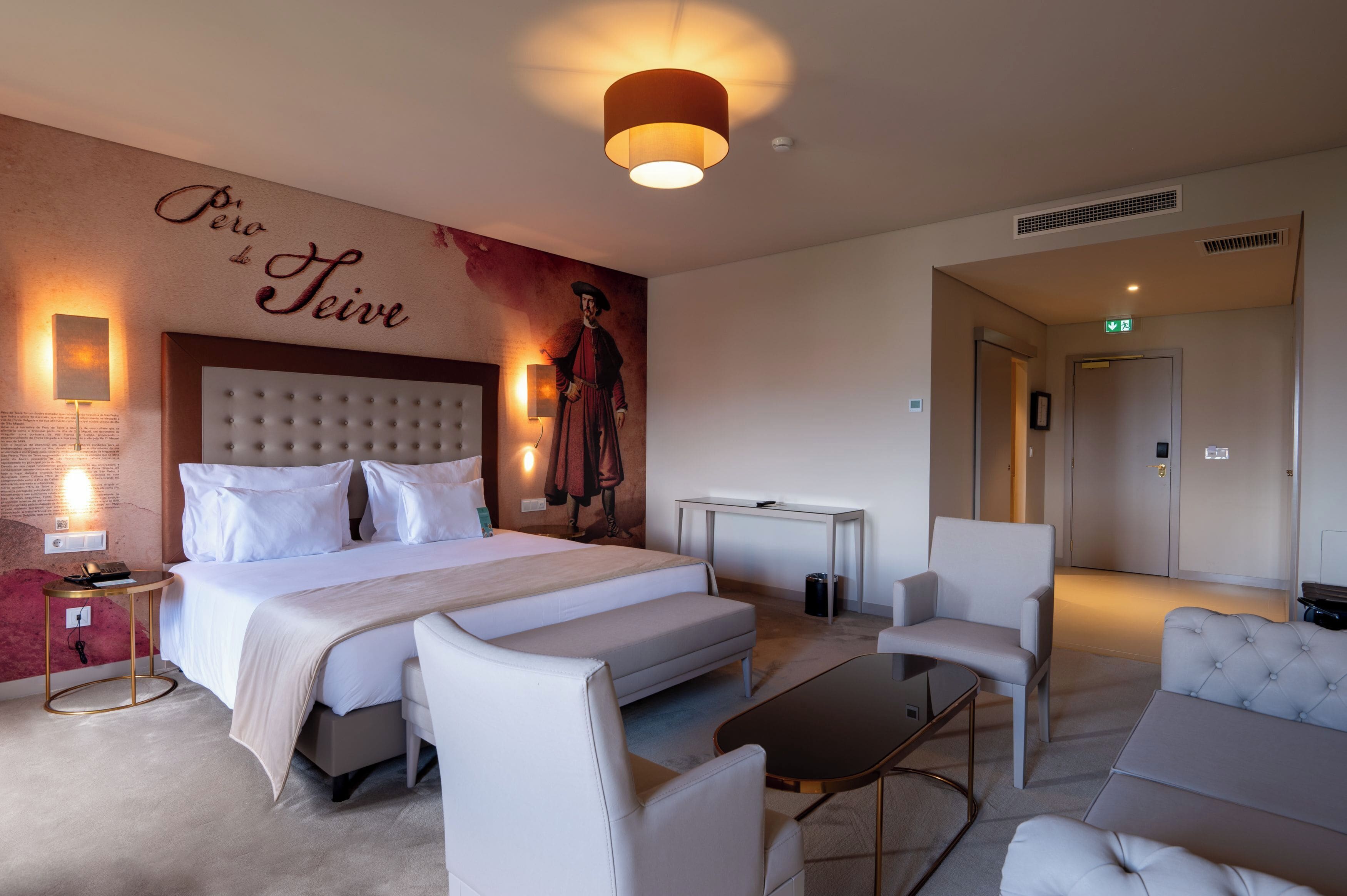 A hotel room with a large bed, a sofa, a coffee table, and a desk. There is a wall with a mural and a sign that reads "Perro de Teive".