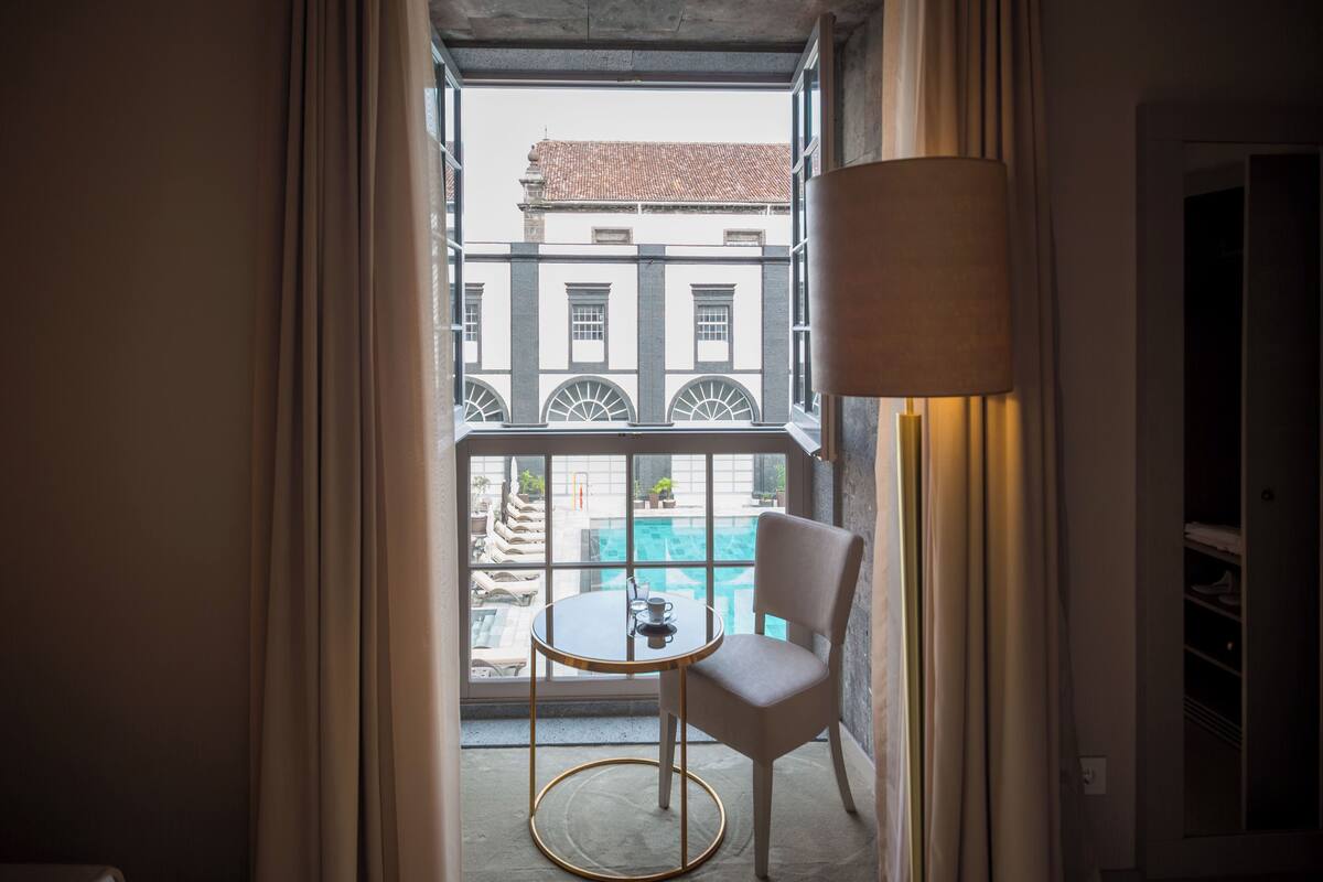 A room with a view of a building through a window, a round table with a chair, and a lamp.