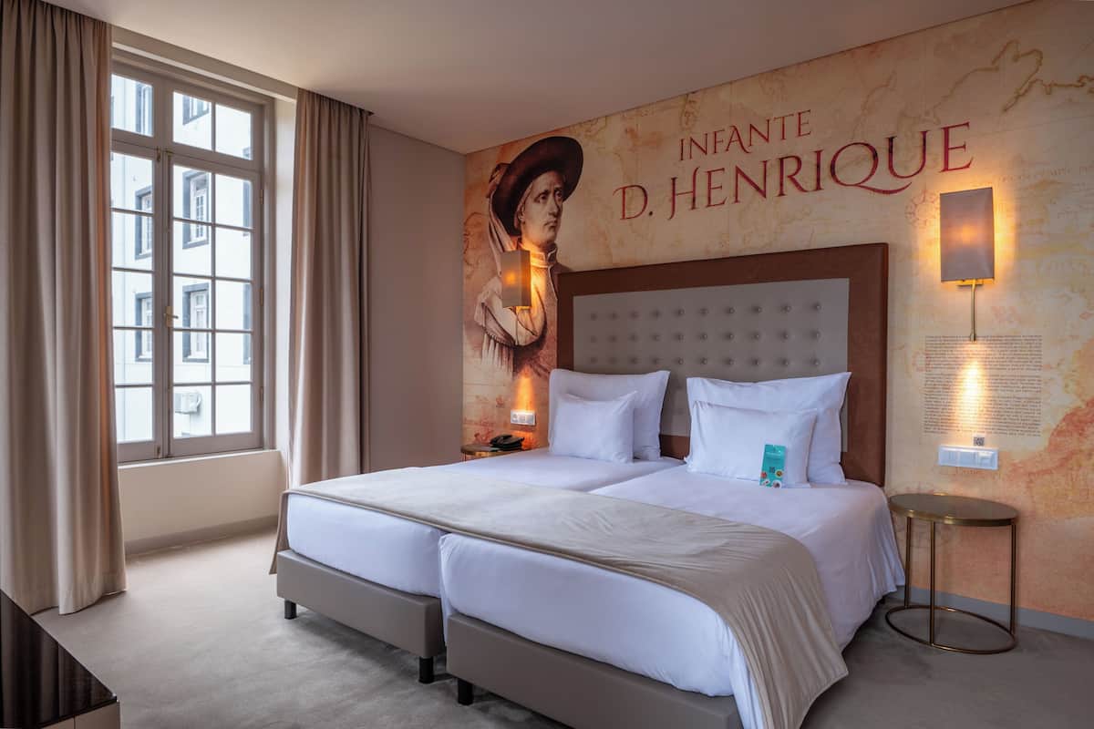 A hotel room with two beds, a large window with curtains, a wall mural, and a round table with a lamp.
